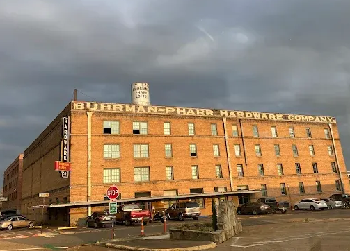 Large brick building: Buhman-Pharr Hardware Company, with cars parked out front, under a cloudy sky.