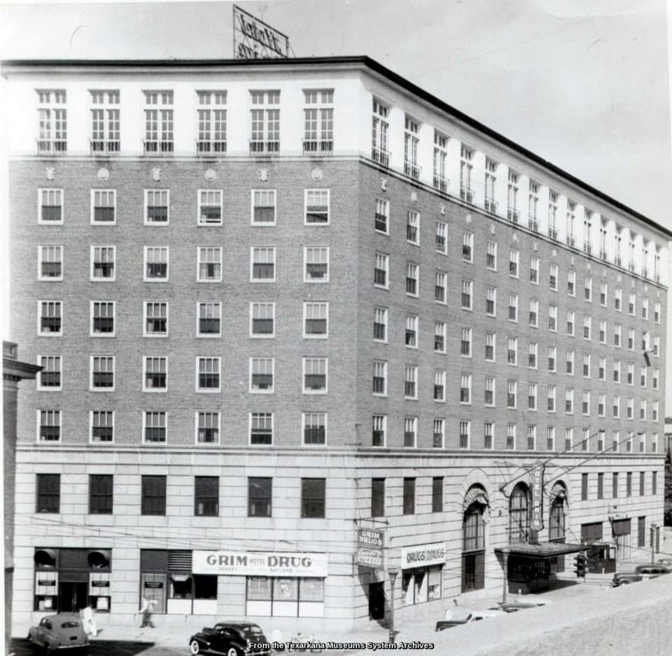 Black and white photo of a tall brick building, possibly a hotel, with many windows. A few cars are parked in front.