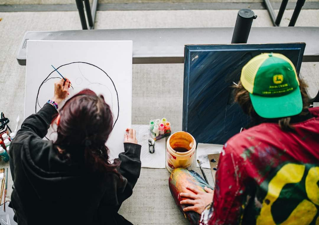 Two people painting at a table; one draws a circle, the other paints a blue square. Paint supplies are visible.