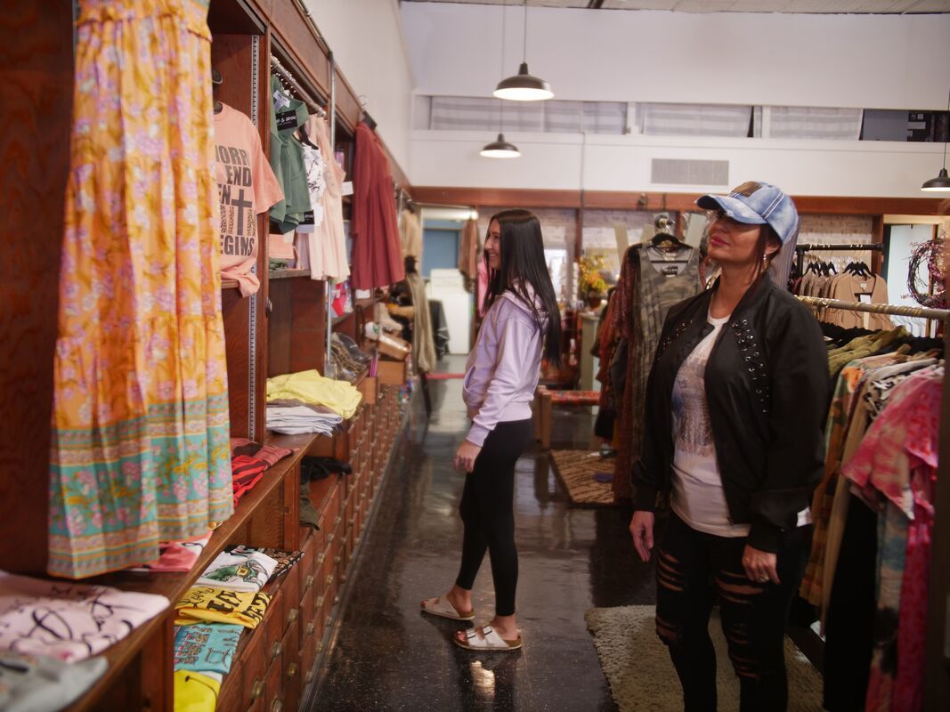 Two women browsing clothes in a vintage clothing store. One in a baseball cap, looking up; the other, a sweatshirt.