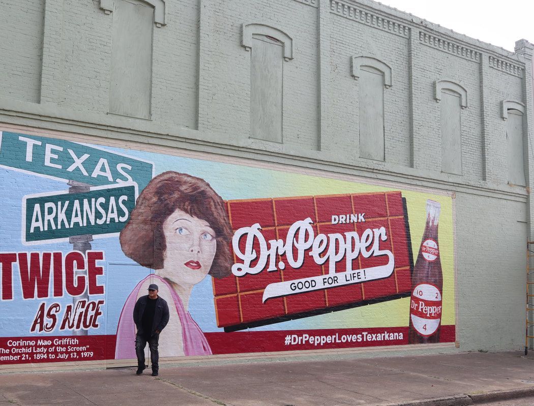 Man stands before a brick wall mural: Dr. Pepper ad with a woman, sign for Texas & Arkansas.