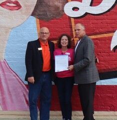 Three people stand in front of a colorful mural. A woman holds a document as two men flank her.