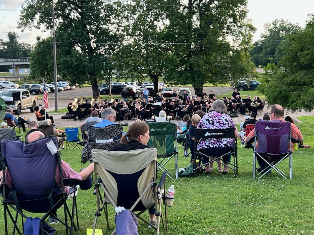 Outdoor band concert with audience in lawn chairs. Band in black uniforms. Green trees and sky.