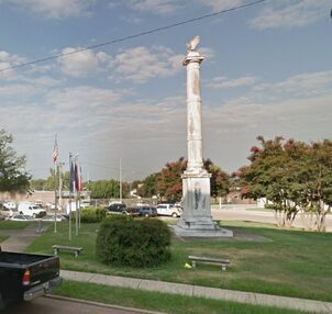 Monument with an eagle on top, surrounded by green grass and trees, small benches, and American flags.