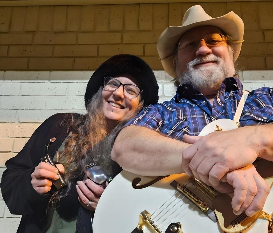 Couple smiling, one holding a white guitar, the other a harmonica, against a brick wall.