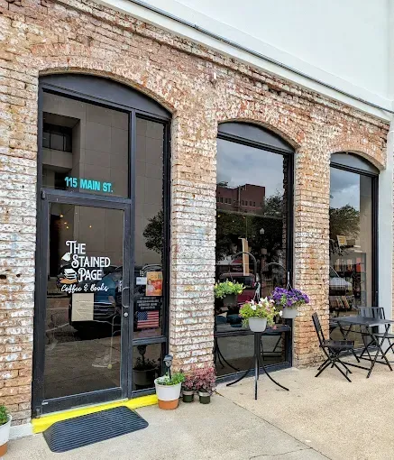 The Stained Page bookstore entrance: arched windows, brick facade, door with sign, outdoor seating, flowers.