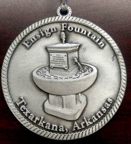 Medallion depicting the Ensign Fountain in Texarkana, Arkansas. Silver-colored metal, with a raised fountain design and text.