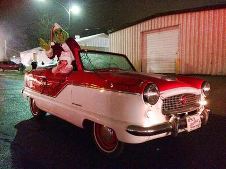 Grinch in a Santa suit waves from a red and white vintage car at night.
