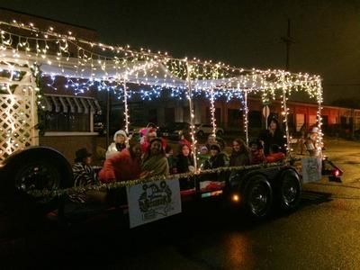 Float in a Christmas parade, lit with white lights, carrying people at night.