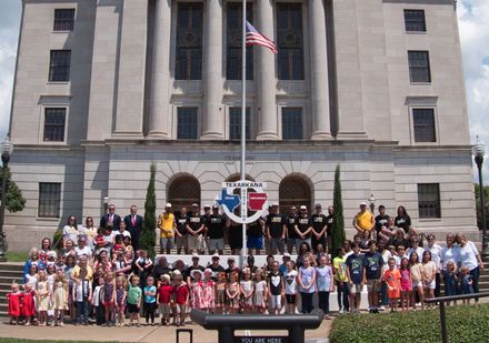 Large group of people posing in front of a building with American flag. A large logo is in the center.