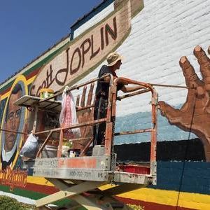 Artist painting a Scott Joplin mural from a lift. The mural has a portrait, the name “Scott Joplin,” and a hand.