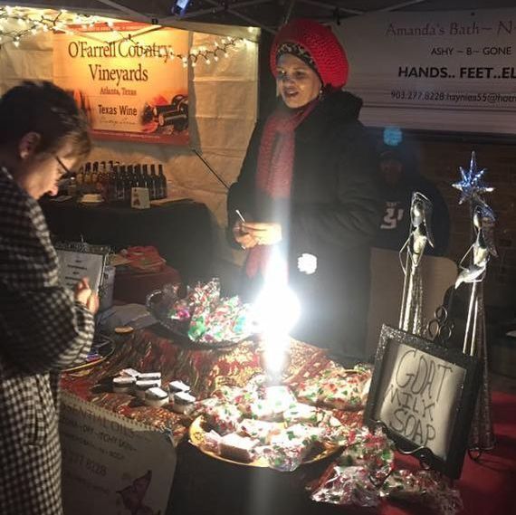 A woman stands behind a table with baked goods at a night market, lit by string lights and a bright light.