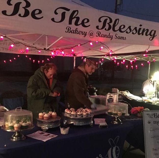 Two men at a bakery stand under a tent with pink lights. Cupcakes and baked goods are displayed on the table.