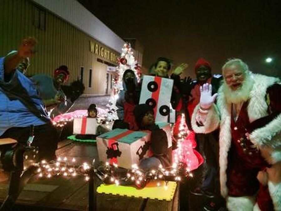 A float in a Christmas parade with people waving, a Santa Claus, and gift box decorations with lights.