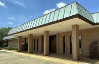 Exterior view of a beige building with a flat, green-roofed awning supported by columns. Sunny day.