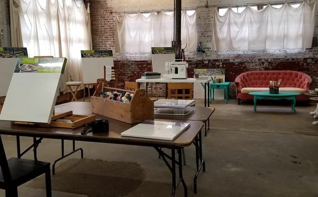 Art studio with easels, paint supplies, and a red sofa. Brick wall, white curtains.
