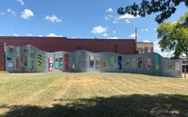 A long, wavy, gray wall with colorful panels in front of a brick building under a blue sky.