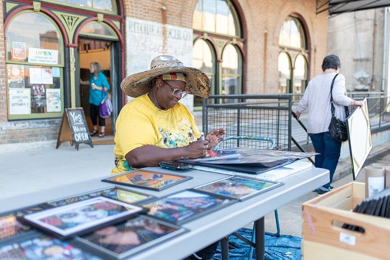 Woman at a table selling artwork outside a brick building. She wears a hat and yellow shirt, working on a piece.