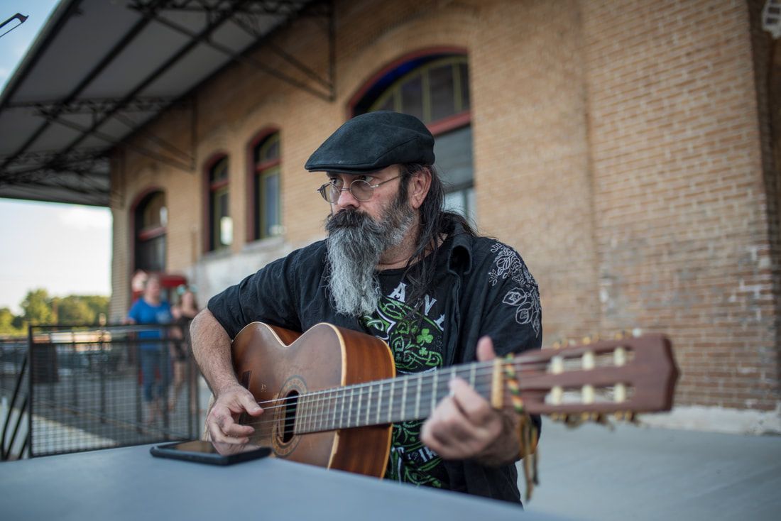 Man with a long beard and glasses playing an acoustic guitar outdoors. He's wearing a cap and black shirt.