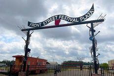 Sign for Front Street Festival Plaza, arched with text, decorated with metal art. Red caboose visible.