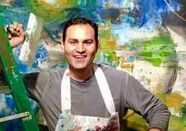 Man wearing apron smiles in front of a colorful abstract painting.