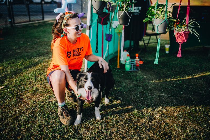 Woman in orange shirt pets a black and white dog in grassy area near a plant display.