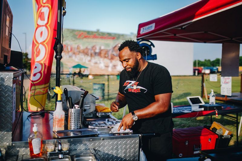 Man cooking food at a food stand with a red canopy, Budweiser mural in the background.