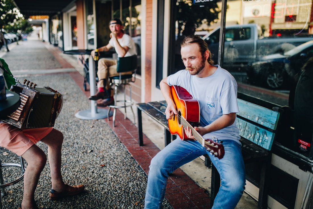 Street musicians playing guitar and accordion outside a shop. A man sits nearby.
