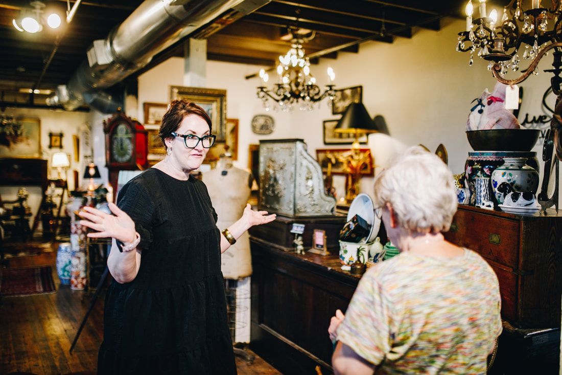 Woman gestures, talking to another in an antique shop filled with furniture and decor.
