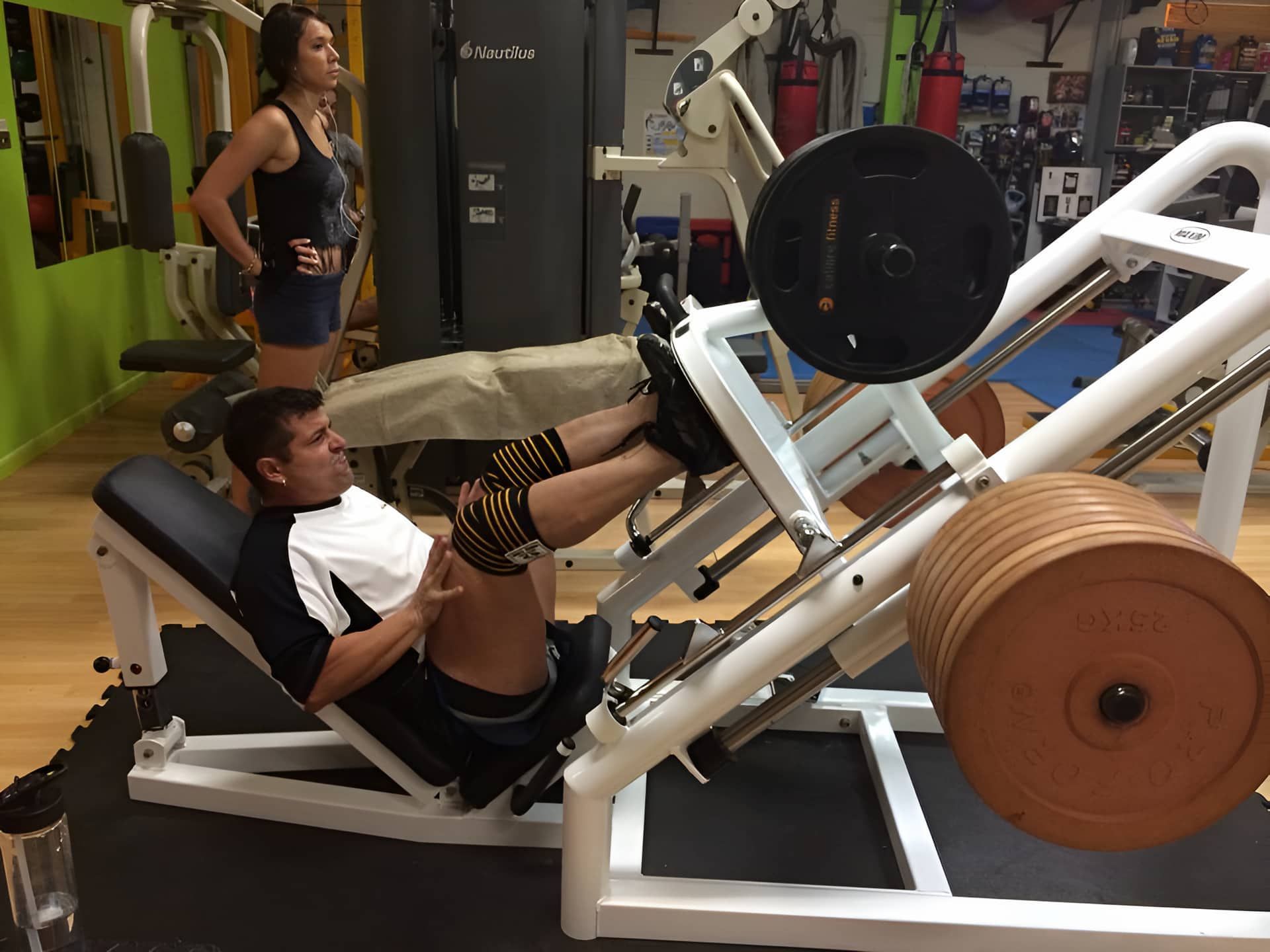 A Man Is Using A Leg Press Machine In A Gym While A Woman Watches — Maxenergy Fitness In Smithfield, QLD