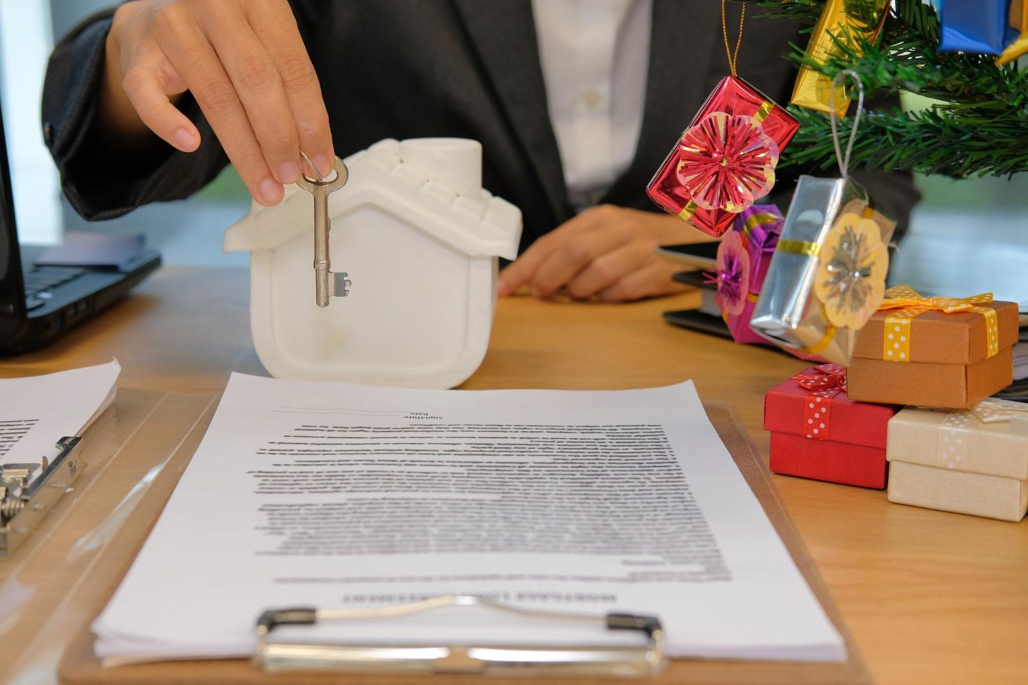 Person holding a key over a small white house model on a desk with a signed document, gifts, and a Christmas tree