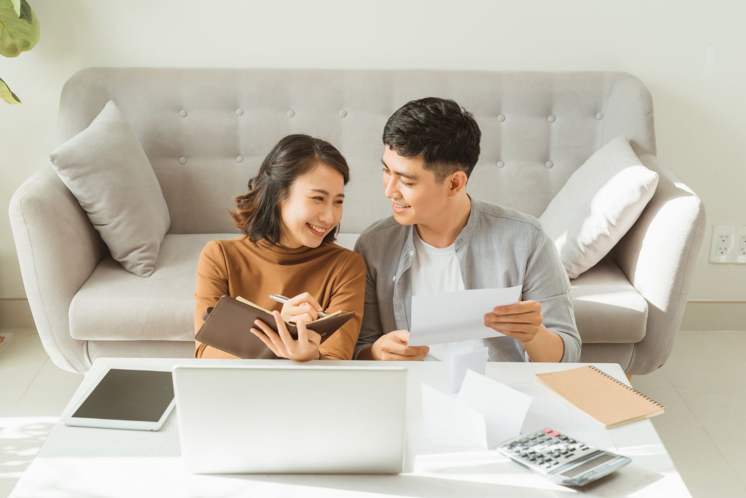 Couple reviews finances, smiling. Woman writes in notebook, man holds bills. White living room, sunny.
