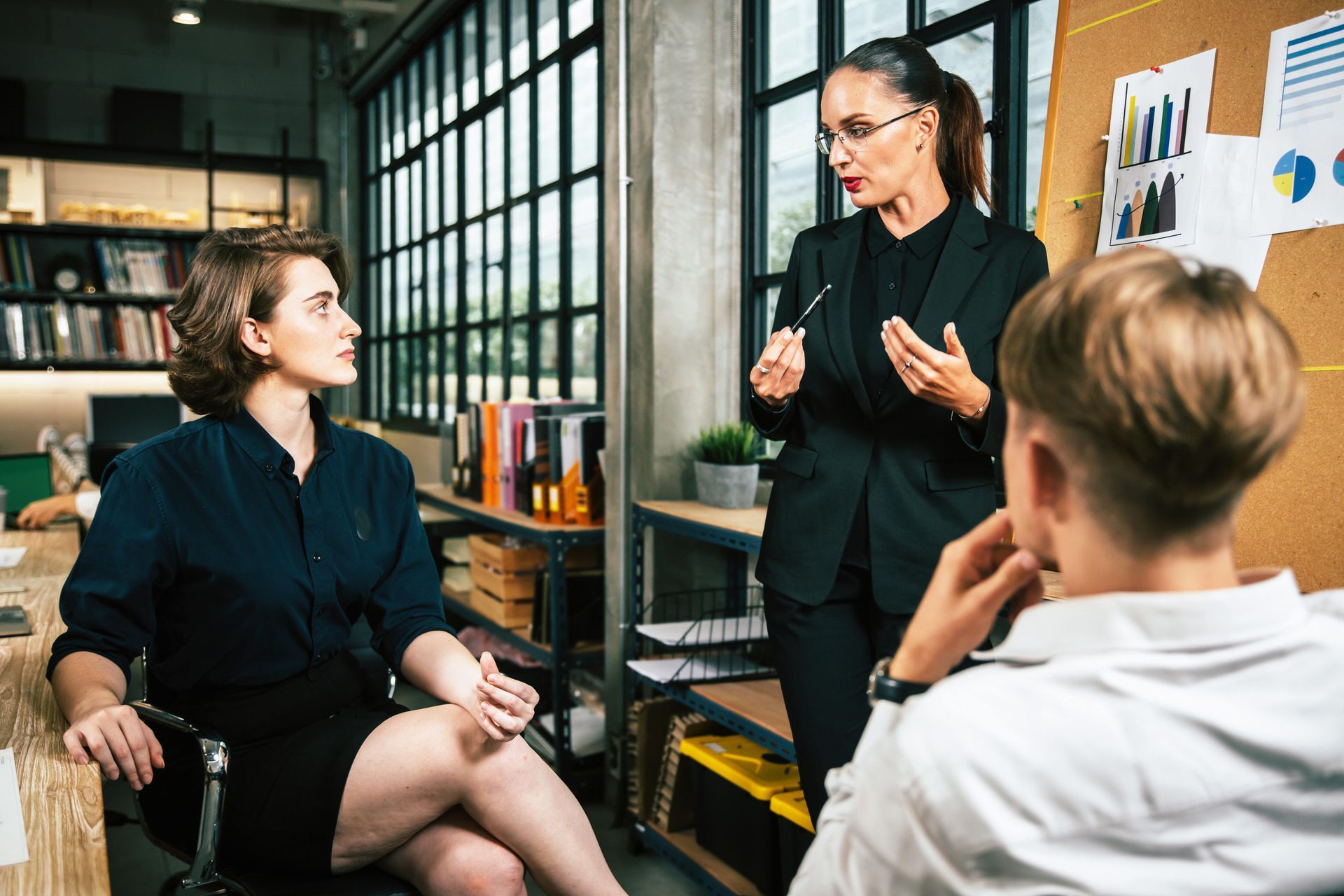 Businesswoman in a suit speaks to two colleagues in an office setting; one seated, one listening