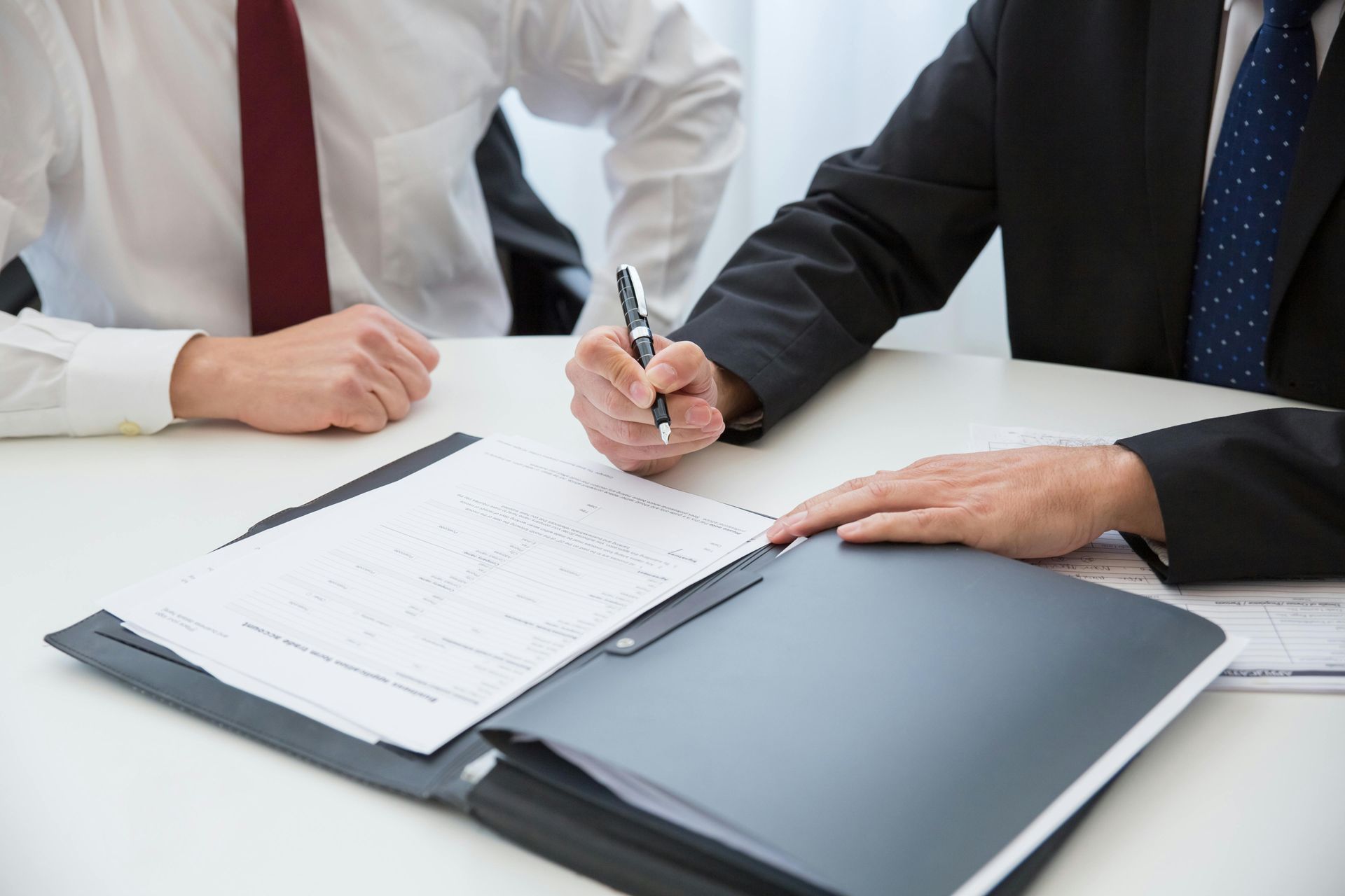 Two men in suits at a table; one signs a document, the other watches.
