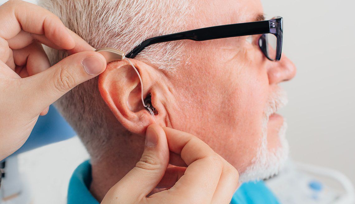 Hands adjust an elderly man's hearing aid behind his ear as he wears black glasses.