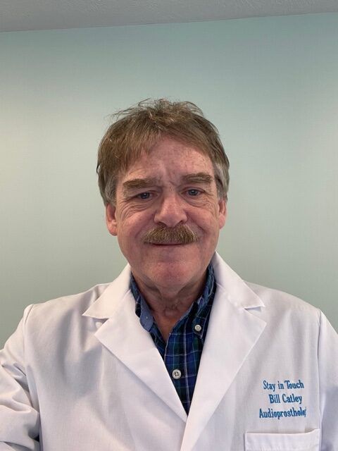 Portrait of a man in a white lab coat and blue shirt, standing against a plain light wall.