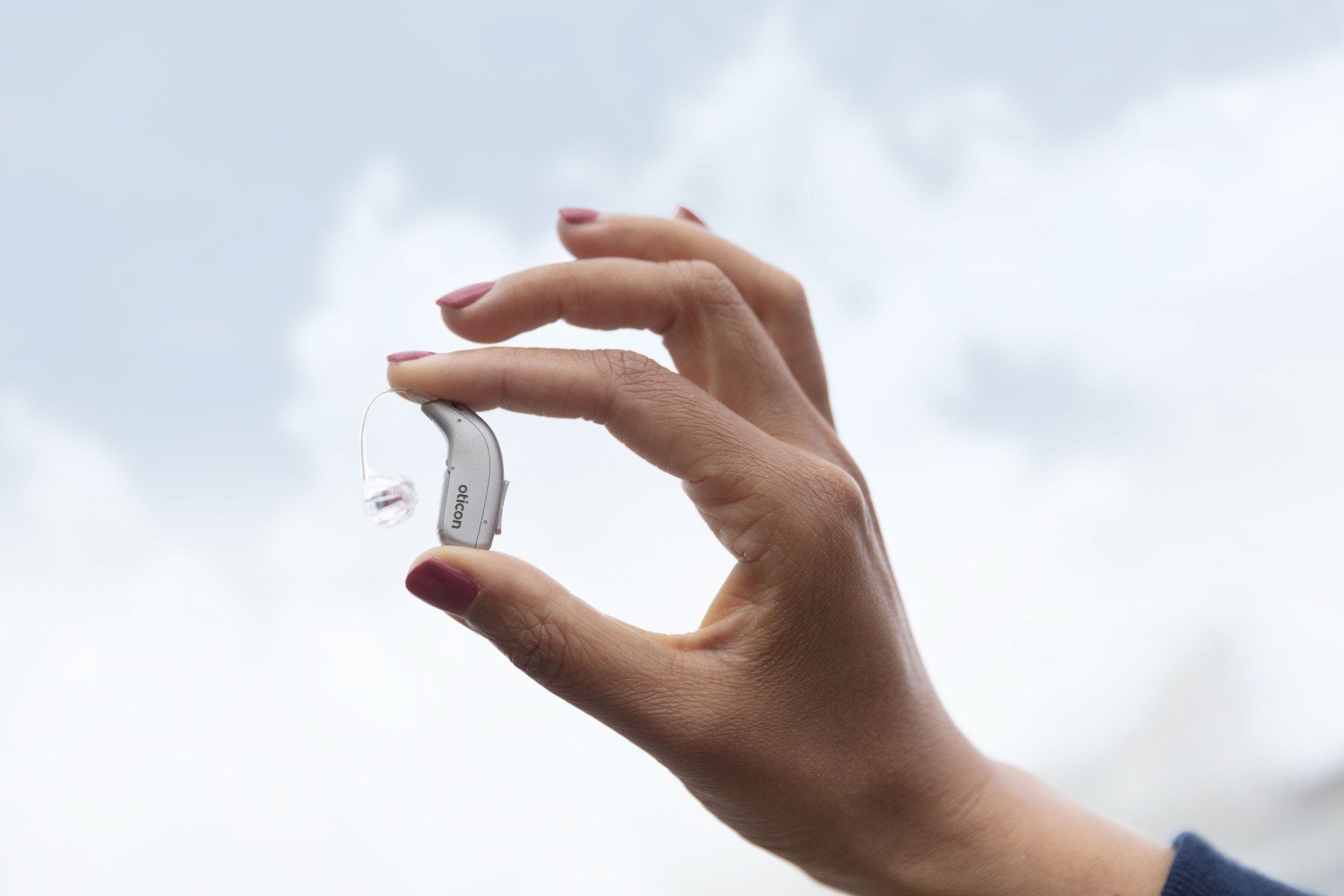 Hand holding a small translucent hearing aid against a cloudy sky