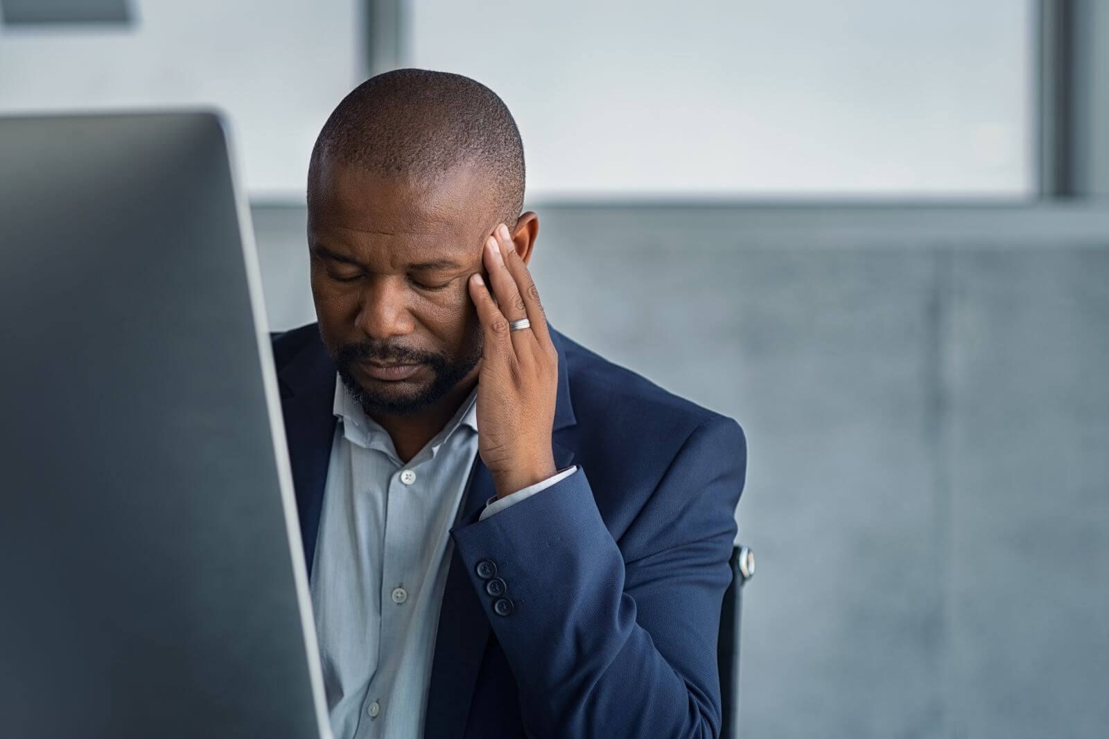 A man in a blue suit sits at a desk, holding his temple beside a laptop, looking stressed.