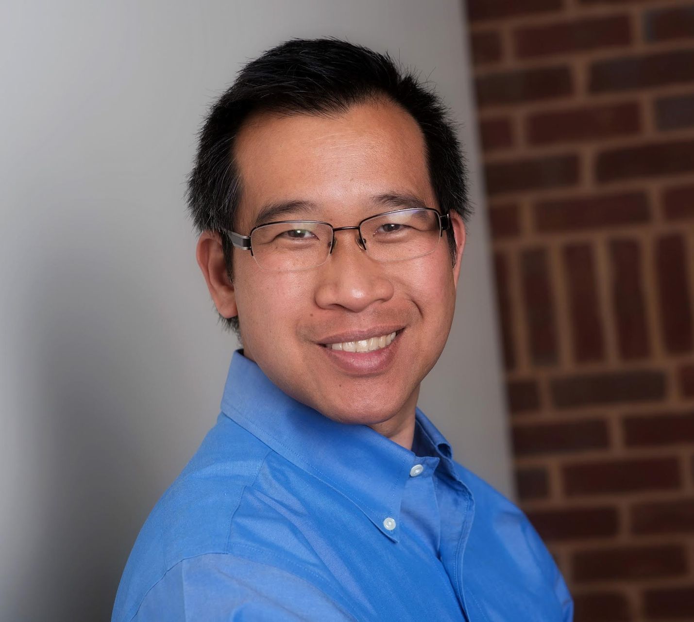 A man wearing glasses and a blue shirt is smiling in front of a brick wall.