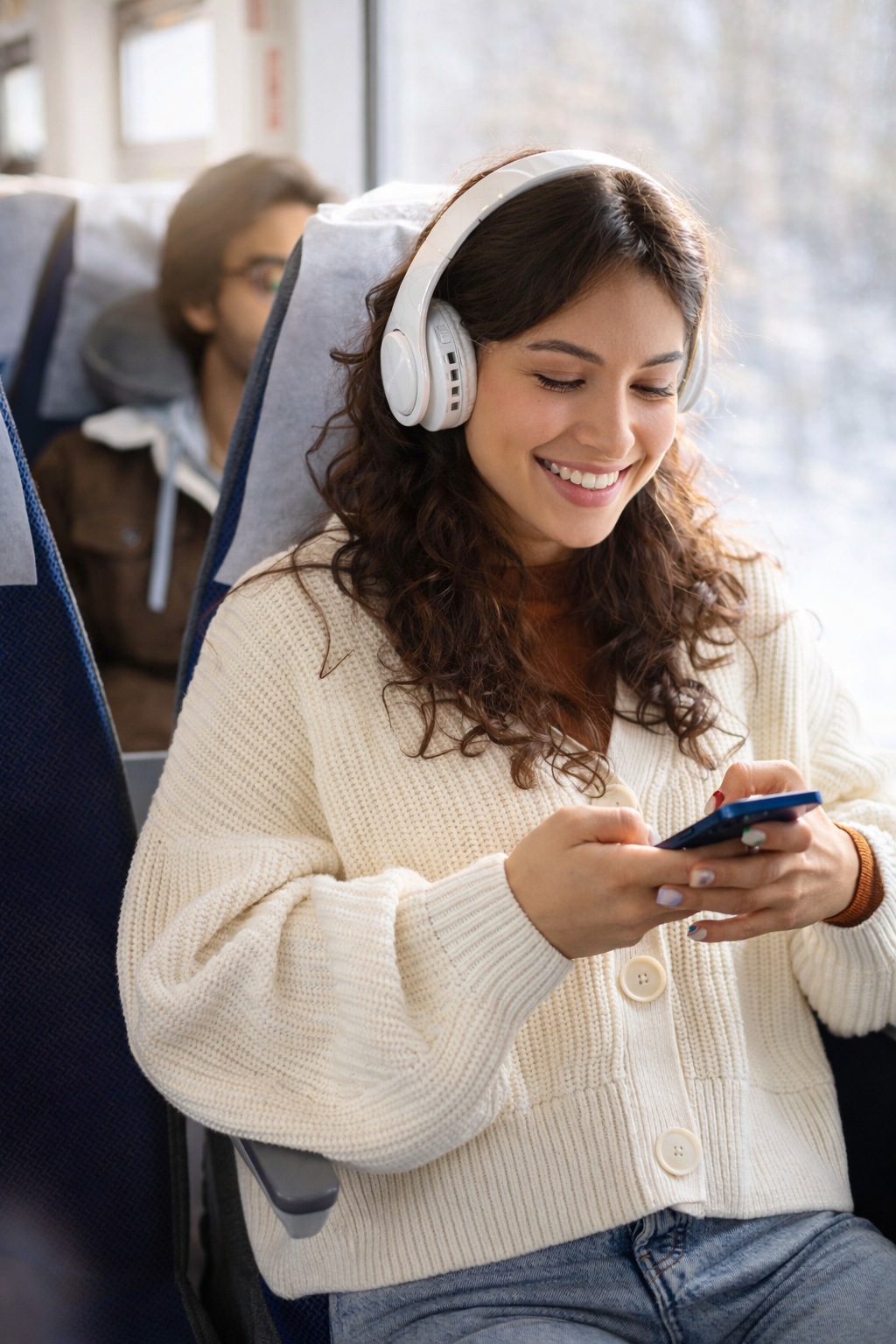 Mujer sonriente con suéter color crema y auriculares blancos usando un teléfono inteligente en un tren o autobús.