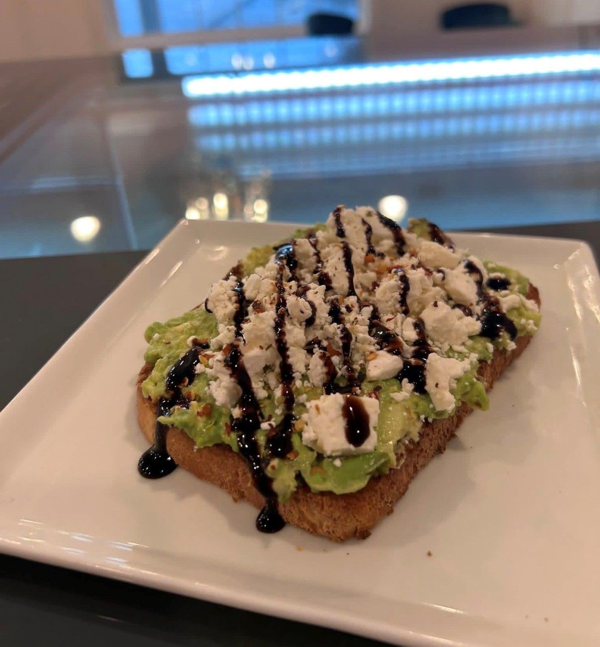 Avocado toast with drizzled balsamic and crumbled cheese on a white plate in a café setting