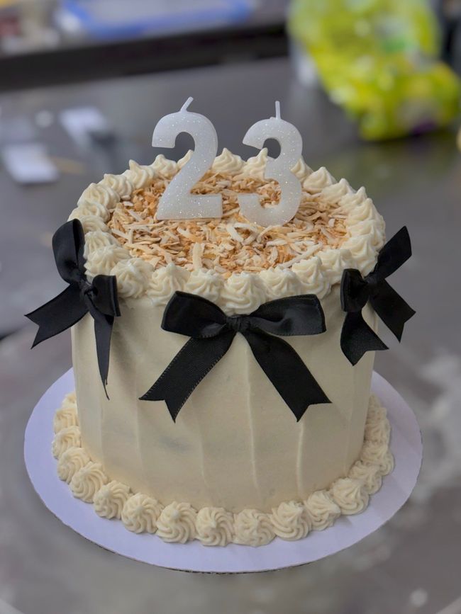 White frosted birthday cake with black ribbons and a silver “23” topper on a table