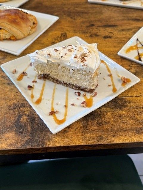 Slice of cheesecake on a white plate with caramel drizzle and crumbs on a wooden table.