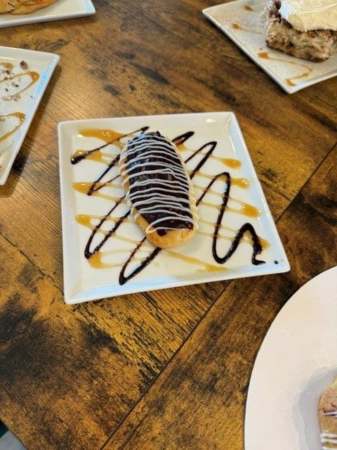 Chocolate-drizzled rolled dessert on a white plate atop a wooden table, with other dessert plates nearby