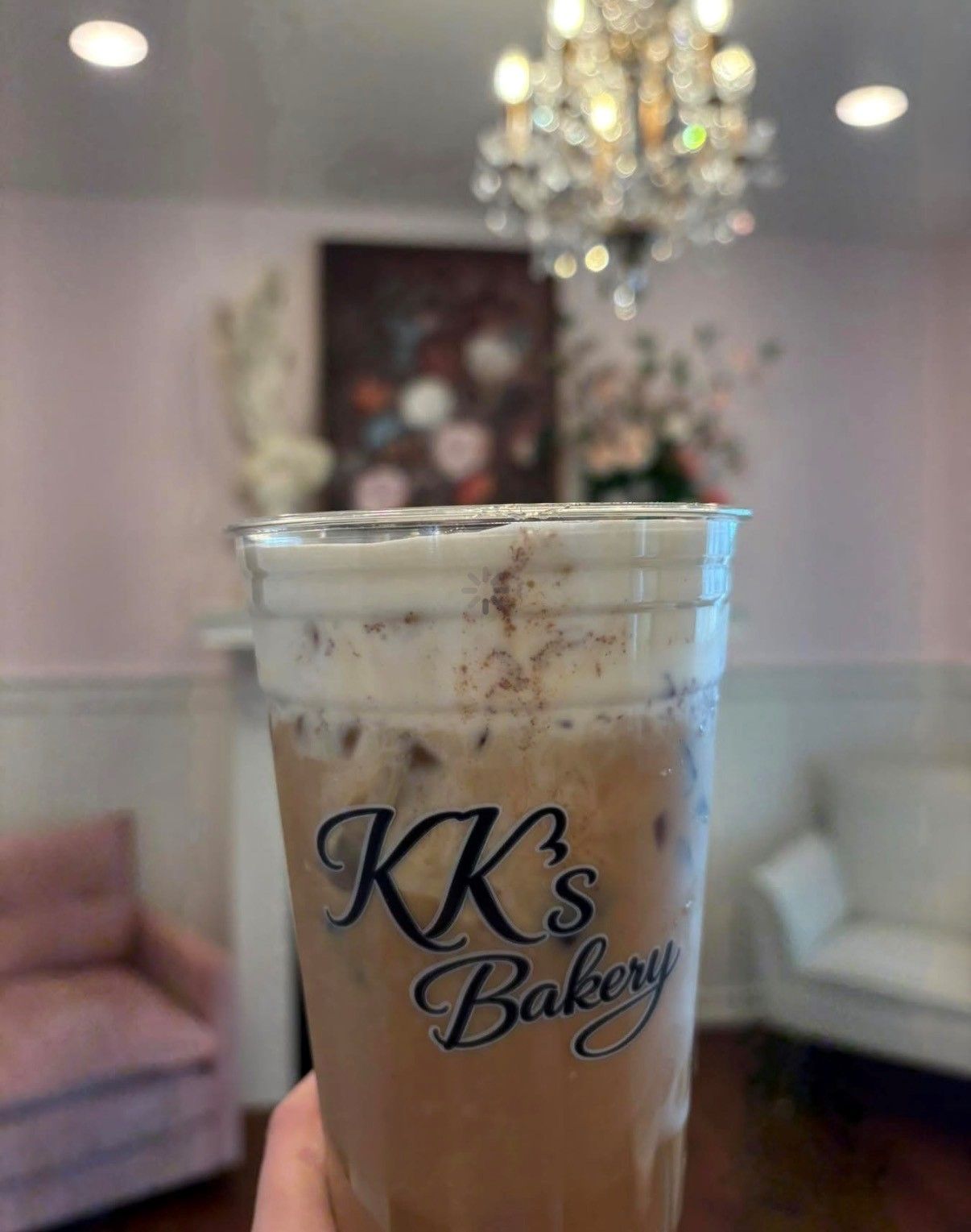 Iced coffee in a clear cup labeled “KK’s Bakery” against a pastel café interior