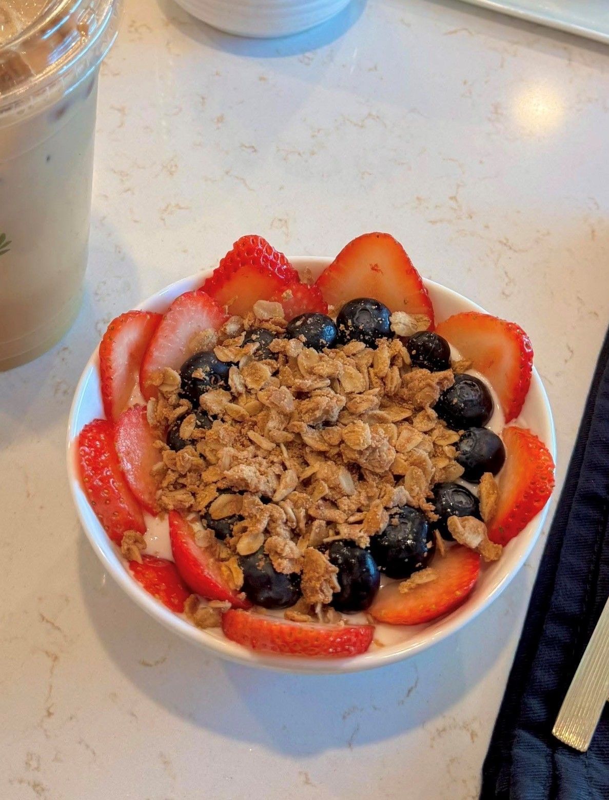 Bowl of granola topped with strawberries and blueberries on a table, with a drink beside it