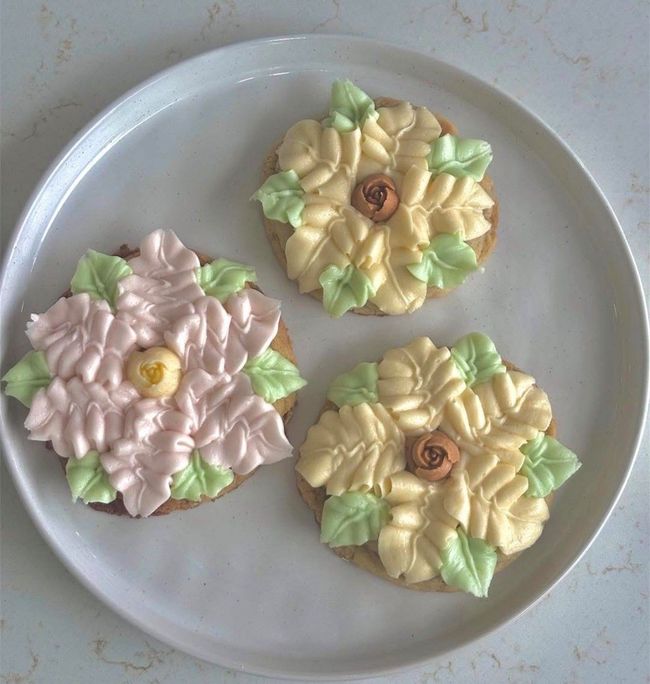 Three frosted flower-shaped cookies on a white plate, one pink and two yellow with green leaves.