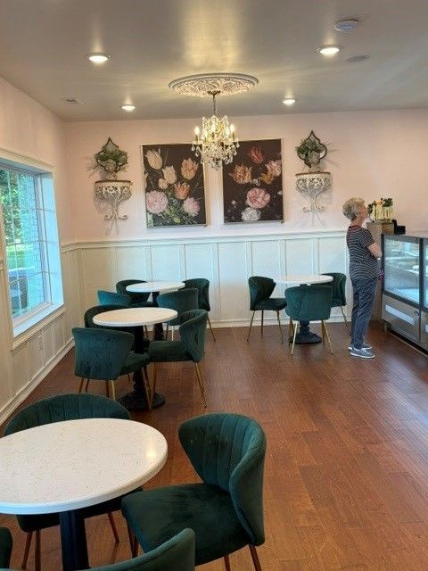 Bright café with green velvet chairs, round tables, wall art, and a woman near the counter.