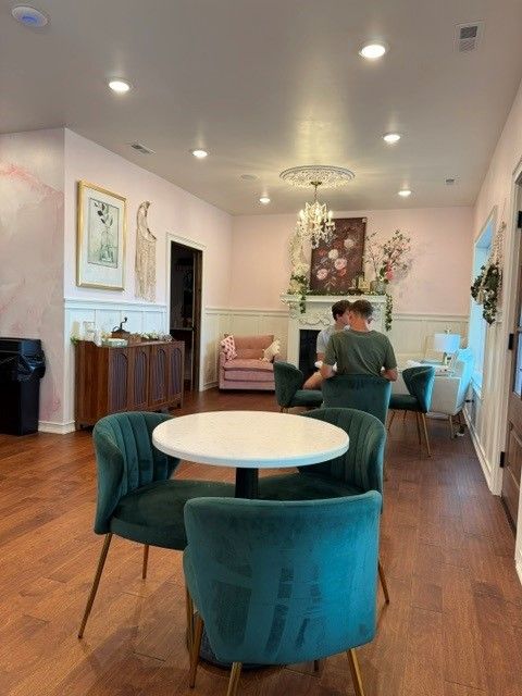 Elegant lounge with teal chairs, round table, and two people talking near a fireplace and floral decor.