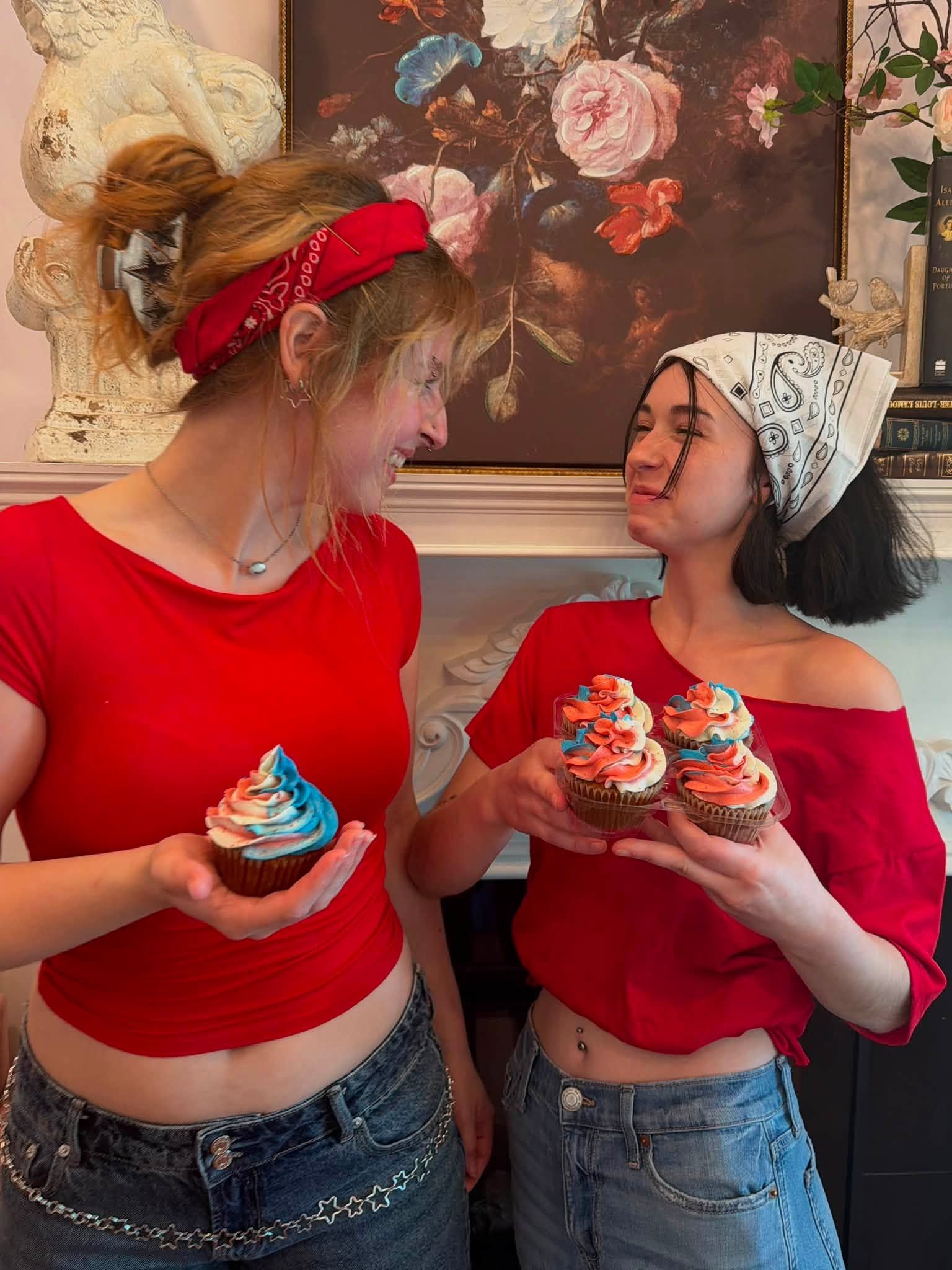 Two women in red tops and jeans hold colorful cupcakes and smile at each other indoors.